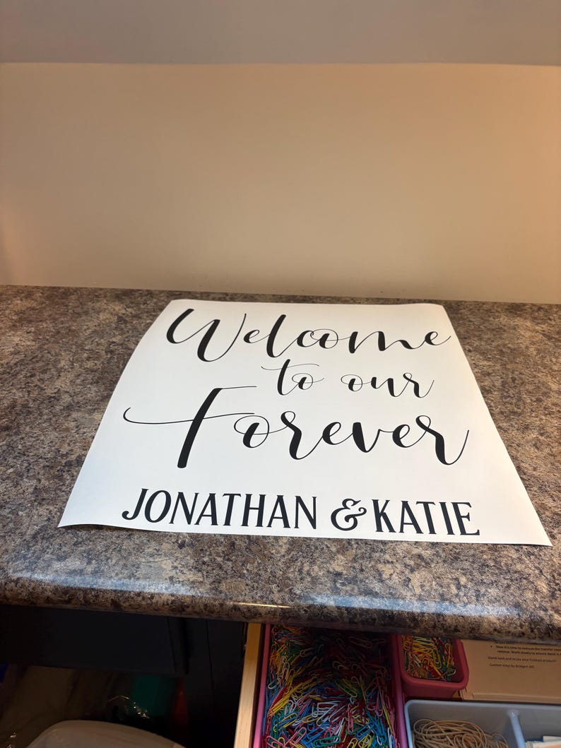 May include: A white rectangular sign featuring black text that reads Welcome to our Forever and JONATHAN & KATIE. The sign uses a combination of a decorative script font for the first three words and a bold sans-serif font for the names.