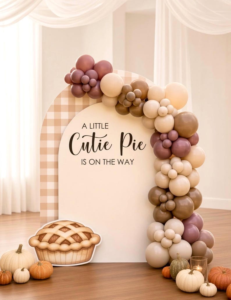 May include: An arched backdrop with a beige gingham pattern and the text A LITTLE Cutie Pie IS ON THE WAY. A balloon garland in shades of cream, tan, and mauve curves over the arch. A cutout of a pie sits at the base with assorted pumpkins.