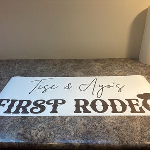 以下が含まれることがあります： 黒い文字で「Tise & Ayo's FIRST RODEO」と書かれた白いビニール製デカール。FIRSTの文字は太字のスタイリッシュなフォントで、RODEOの最後のOの文字にはカウボーイハットのアイコンが組み込まれています。
