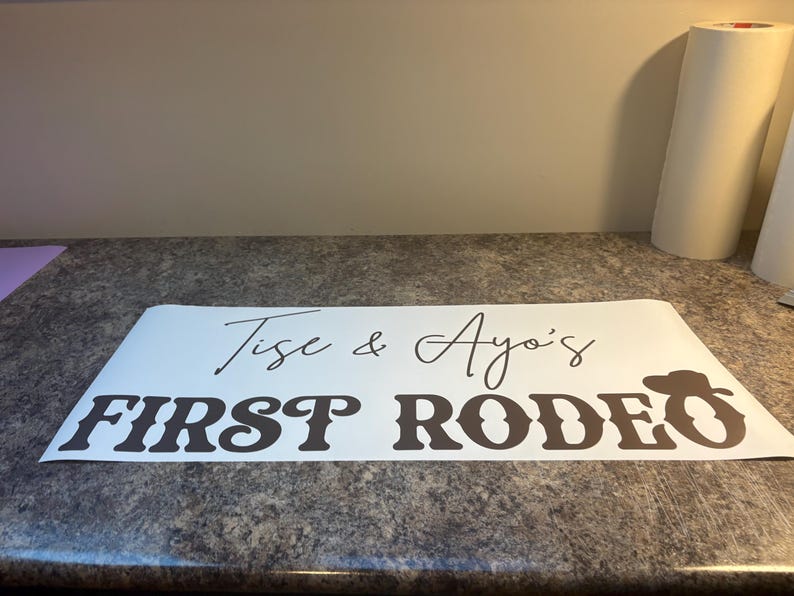 以下が含まれることがあります： 黒い文字で「Tise & Ayo's FIRST RODEO」と書かれた白いビニール製デカール。FIRSTの文字は太字のスタイリッシュなフォントで、RODEOの最後のOの文字にはカウボーイハットのアイコンが組み込まれています。