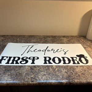 以下が含まれることがあります： 黒い文字で「Theodore's FIRST RODEO」と書かれた白いビニール製デカール。RODEOの「O」の文字の上には、小さな黒いカウボーイハットのアイコンが配置されています。デカールは平らな長方形の台紙に貼られています。