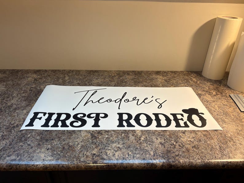 以下が含まれることがあります： 黒い文字で「Theodore's FIRST RODEO」と書かれた白いビニール製デカール。RODEOの「O」の文字の上には、小さな黒いカウボーイハットのアイコンが配置されています。デカールは平らな長方形の台紙に貼られています。