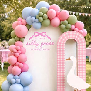May include: A whimsical party decoration featuring a white arch adorned with pastel-colored balloons in pink, blue, and green. A pink and white checkered archway frames a white goose cutout. The text "A little silly goose is almost loose" is displayed on the arch.