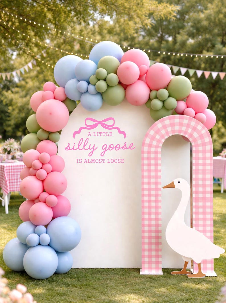 May include: A whimsical party decoration featuring a white arch adorned with pastel-colored balloons in pink, blue, and green. A pink and white checkered archway frames a white goose cutout. The text "A little silly goose is almost loose" is displayed on the arch.