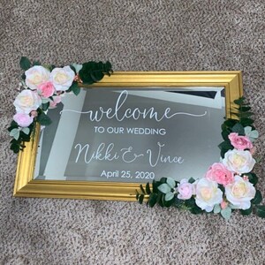 Może przedstawiać: Prostokątne lustro w złotej ramie z białym napisem welcome TO OUR WEDDING Nikki & Vince April 25, 2020. Lustro jest ozdobione sztucznymi r&oacute;żowymi i białymi r&oacute;żami oraz zielonymi liśćmi w lewym g&oacute;rnym i prawym dolnym rogu.