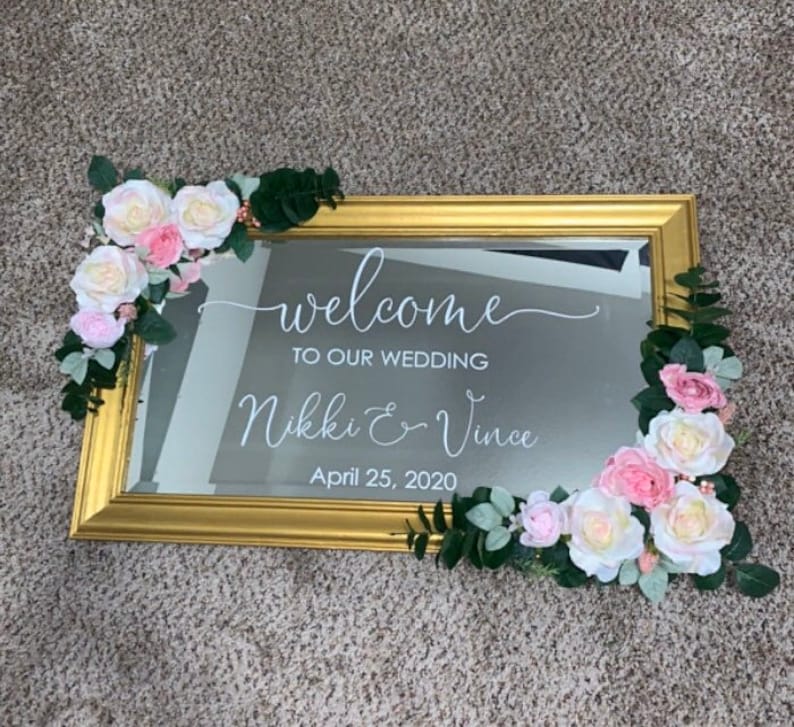 Może przedstawiać: Prostokątne lustro w złotej ramie z białym napisem welcome TO OUR WEDDING Nikki & Vince April 25, 2020. Lustro jest ozdobione sztucznymi r&oacute;żowymi i białymi r&oacute;żami oraz zielonymi liśćmi w lewym g&oacute;rnym i prawym dolnym rogu.