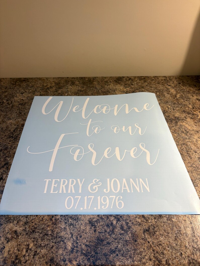 May include: A light blue square decal featuring white text that reads Welcome to our Forever, followed by the names TERRY & JOANN and the date 07.17.1976 in a clean, sans-serif font. The decal has a smooth, matte finish and is ready for application.