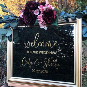 Może przedstawiać: Prostokątne lustro w złotej ramie ze złotym napisem welcome TO OUR WEDDING Cody & Shelby 02.29.2020. Lustro ozdobione jest bukietem ciemnoczerwonych i jasnor&oacute;żowych sztucznych kwiat&oacute;w oraz zielonymi liśćmi eukaliptusa.