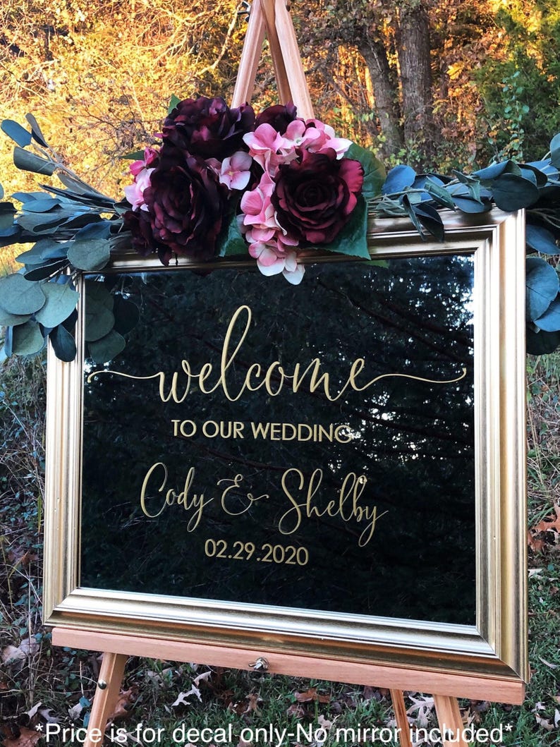 Może przedstawiać: Prostokątne lustro w złotej ramie ze złotym napisem welcome TO OUR WEDDING Cody & Shelby 02.29.2020. Lustro ozdobione jest bukietem ciemnoczerwonych i jasnor&oacute;żowych sztucznych kwiat&oacute;w oraz zielonymi liśćmi eukaliptusa.