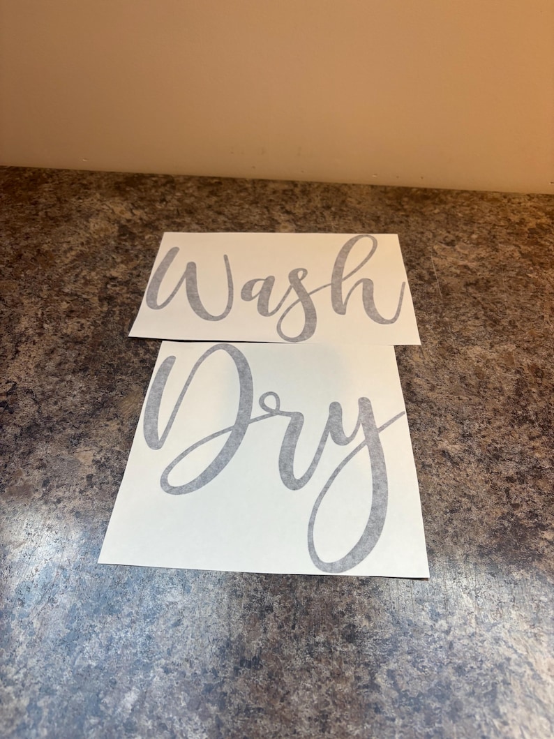 Puede incluir: Dos calcoman&iacute;as rectangulares blancas con texto cursivo gris. La calcoman&iacute;a superior dice "Wash", y la calcoman&iacute;a inferior dice "Dry". Las calcoman&iacute;as est&aacute;n colocadas sobre una superficie oscura y texturizada.