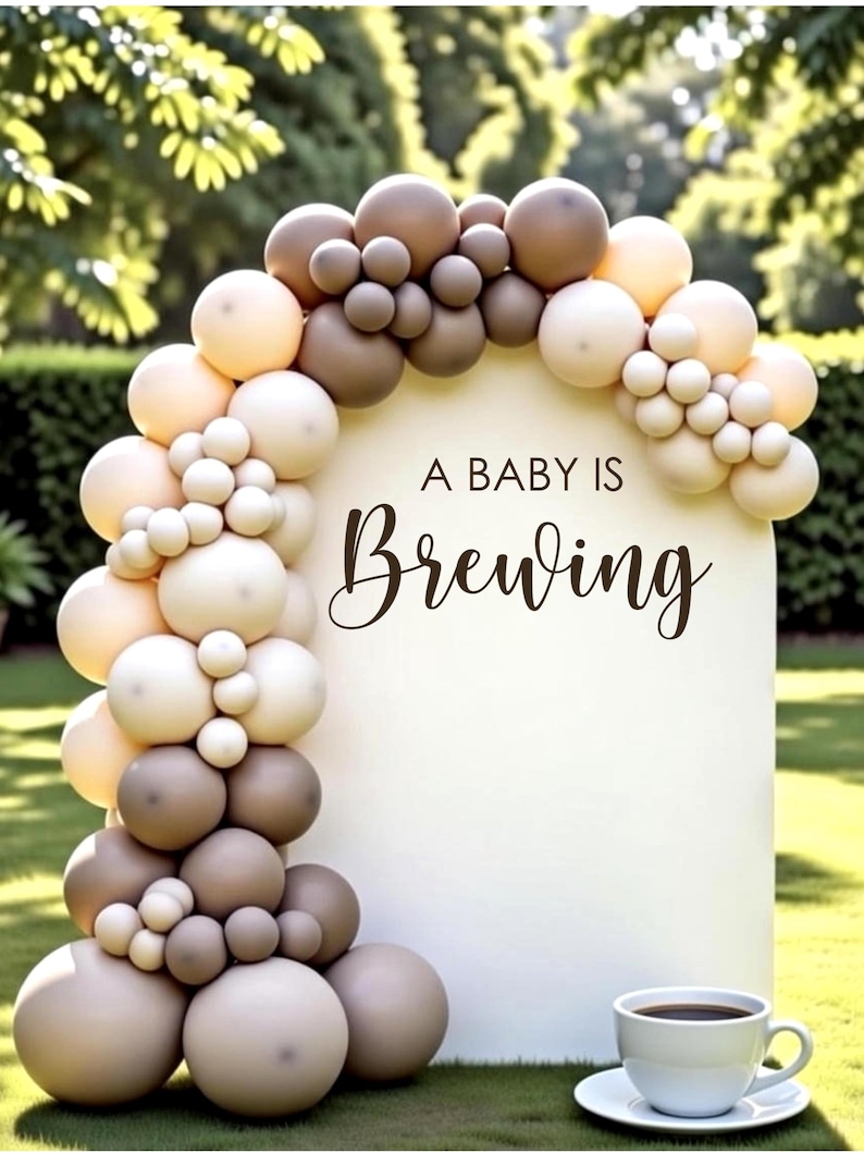 May include: A white sign with the text "A BABY IS Brewing" is surrounded by a balloon arch in shades of brown and cream. A white teacup and saucer sit on the grass in front of the sign. The background is a green lawn and foliage.