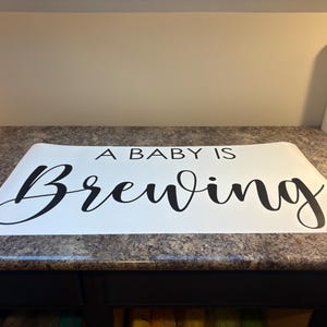 May include: A white rectangular sign with the phrase "A BABY IS Brewing" in black cursive font. The sign is placed on a speckled countertop. A roll of white material is visible in the background.