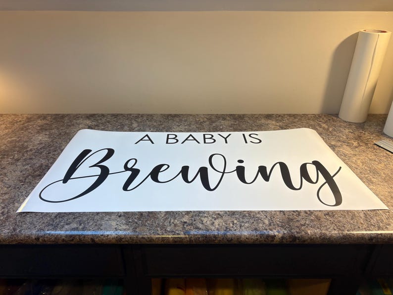 May include: A white rectangular sign with the phrase "A BABY IS Brewing" in black cursive font. The sign is placed on a speckled countertop. A roll of white material is visible in the background.