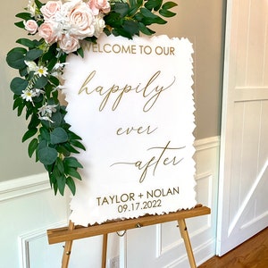 Peut inclure: Une pancarte en acrylique blanche avec des lettres dor&eacute;es sur un chevalet. La pancarte indique "Bienvenue &agrave; notre bonheur pour toujours Taylor + Nolan 09.17.2022". La pancarte est d&eacute;cor&eacute;e d'une guirlande de fleurs blanches et roses et de verdure.