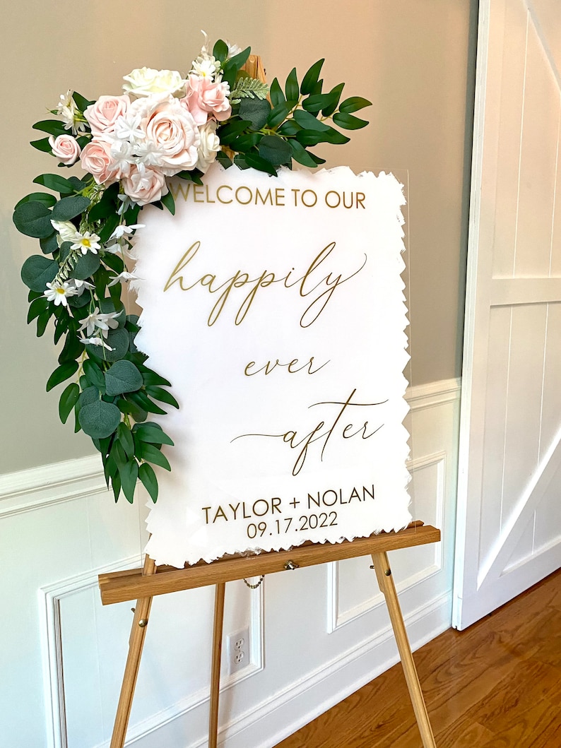 Peut inclure: Une pancarte en acrylique blanche avec des lettres dor&eacute;es sur un chevalet. La pancarte indique "Bienvenue &agrave; notre bonheur pour toujours Taylor + Nolan 09.17.2022". La pancarte est d&eacute;cor&eacute;e d'une guirlande de fleurs blanches et roses et de verdure.
