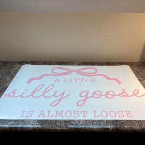 May include: A white sheet with pink text that reads "A LITTLE silly goose IS ALMOST LOOSE". The text is in a playful, cursive font, with a decorative bow above the words "A LITTLE".
