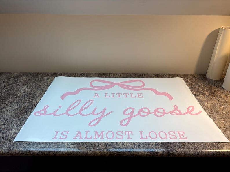 May include: A white sheet with pink text that reads "A LITTLE silly goose IS ALMOST LOOSE". The text is in a playful, cursive font, with a decorative bow above the words "A LITTLE".