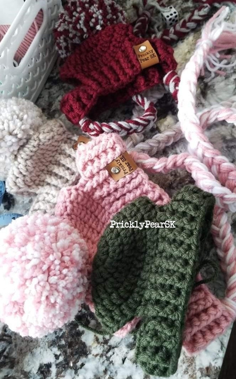 Puede incluir: Una colecci&oacute;n de art&iacute;culos de ganchillo hechos a mano en varios colores. La imagen presenta un art&iacute;culo de ganchillo rojo oscuro con un cord&oacute;n trenzado blanco y rojo, un art&iacute;culo de ganchillo rosa, un art&iacute;culo de ganchillo verde y un pomp&oacute;n rosa y blanco. El texto "PricklyPearSK" es visible.