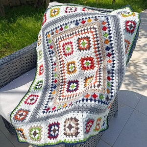 May include: A handmade, colorful crochet blanket draped over a gray wicker chair. The blanket features a square pattern with various colors including red, orange, green, and blue. The border is a mix of gray, white, and a dark blue trim.