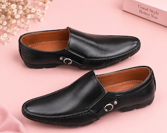 May include: A pair of black slip-on loafers featuring a smooth leather-like finish, a rounded toe, and a decorative silver-toned metal buckle detail on the side. The interior is lined with a tan material, and the soles are black with a textured tread.