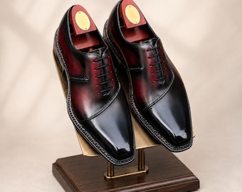 May include: A pair of polished leather oxford shoes featuring a deep burgundy and black gradient finish, black laces, and a sleek pointed toe design. Each shoe is fitted with a wooden shoe tree with a brass-coloured circular metal plate.