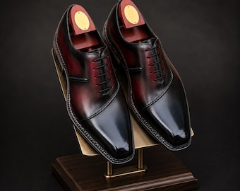 May include: A pair of polished oxfords featuring a deep burgundy and black gradient finish, black laces, and a sleek almond toe. Each shoe contains a wooden shoe tree with a gold-toned metal plate, displayed on a slanted metal stand.