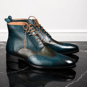May include: A pair of ankle-high boots featuring a two-tone design with deep teal and olive green leather panels. The boots include classic brogue detailing, perforated wingtip patterns, and tan laces threaded through silver-toned metal eyelets.