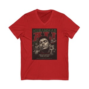May include: Red V-neck t-shirt featuring a graphic of Muhammad Ali. The graphic includes the text "MUHAMMAD ALI" and "The Greatest", along with the quote "FLOAT LIKE A BUTTERFLY STING LIKE A BEE".