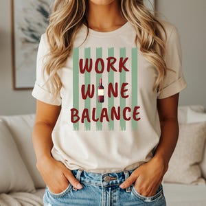 Könnte beinhalten: Ein cremefarbenes Kurzarm-T-Shirt mit einem Grafikdruck aus vertikalen salbeigrünen Streifen. Der Text WORK WINE BALANCE ist in dunkelroten Buchstaben gedruckt, wobei eine kleine Weinflaschenillustration den Buchstaben I in WINE ersetzt.