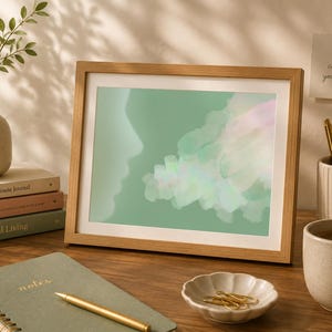 May include: A framed art print with a light wood frame. The artwork features a silhouette of a face and abstract pastel shapes on a green background. The image is on a wooden desk with a notebook, pen, and a cup of tea.