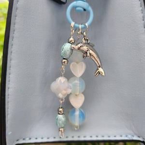 May include: A light blue bag charm featuring a silver-toned dolphin pendant, two white heart-shaped beads, two round opalescent beads, a faceted clover-shaped bead, and small metallic spacer beads, all attached to a light blue plastic ring.