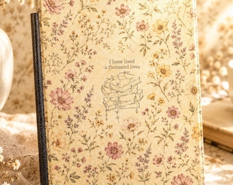 May include: A rectangular e-reader case with a cream background featuring a delicate floral pattern in shades of pink, yellow, and green. The center displays a line drawing of stacked books with the text I have lived a thousand lives.