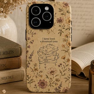 May include: A beige smartphone case featuring a delicate floral pattern in shades of brown, yellow, and pink. The center displays a line art illustration of a stack of books with the text I have lived a thousand lives printed above it.