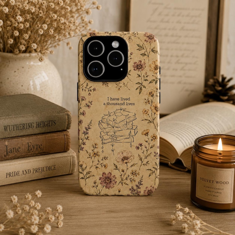 May include: A beige smartphone case featuring a delicate floral pattern in shades of brown, yellow, and pink. The center displays a line art illustration of a stack of books with the text I have lived a thousand lives printed above it.
