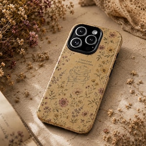 May include: A beige smartphone case featuring a delicate floral pattern in muted tones of yellow, pink, and brown. The design includes a central line-art illustration of a stack of books with the text I have lived a thousand lives above it.