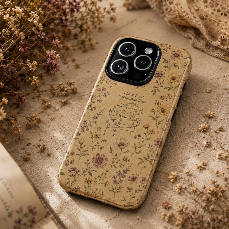 May include: A beige smartphone case featuring a delicate floral pattern in muted tones of yellow, pink, and brown. The design includes a central line-art illustration of a stack of books with the text I have lived a thousand lives above it.