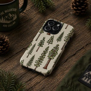May include: A phone case with a cream-colored background featuring a repeating pattern of green and brown pine trees. The phone case is lying on a wooden surface, with a green mug and pine cones in the background.