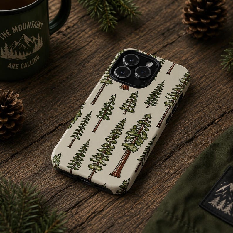 May include: A phone case with a cream-colored background featuring a repeating pattern of green and brown pine trees. The phone case is lying on a wooden surface, with a green mug and pine cones in the background.