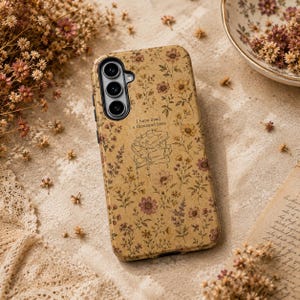 May include: A tan phone case featuring a repeating pattern of small brown and yellow wildflowers. In the center, a line art illustration shows a stack of books with the text I have lived a thousand lives printed above the books.