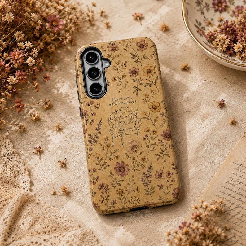 May include: A tan phone case featuring a repeating pattern of small brown and yellow wildflowers. In the center, a line art illustration shows a stack of books with the text I have lived a thousand lives printed above the books.