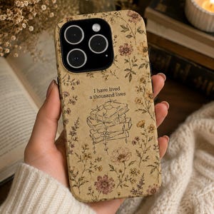 May include: A beige smartphone case featuring a vintage-style floral pattern in muted tones of brown, yellow, and pink. The center displays a line-art illustration of a stack of books with vines and the text, I have lived a thousand lives.