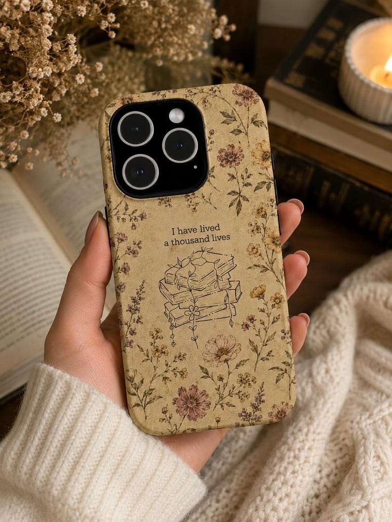 May include: A beige smartphone case featuring a vintage-style floral pattern in muted tones of brown, yellow, and pink. The center displays a line-art illustration of a stack of books with vines and the text, I have lived a thousand lives.