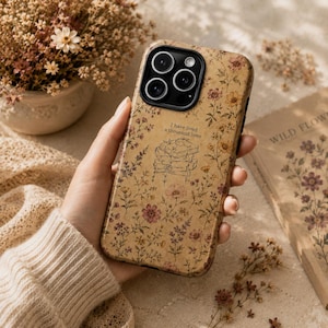 May include: A phone case with a tan background featuring a pattern of small, muted-colored wildflowers and a central line-art illustration of a stack of books with the text, I have lived a thousand lives, printed above the books in black ink.