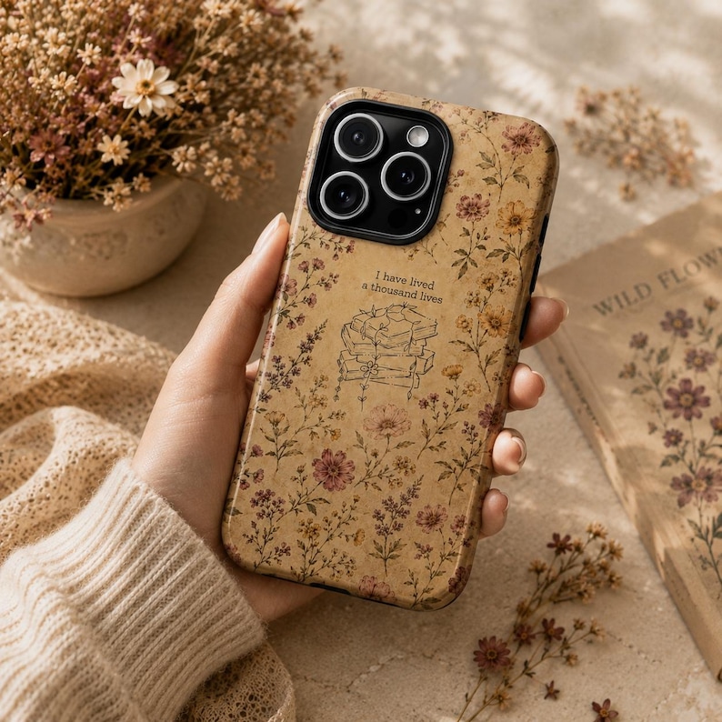 May include: A phone case with a tan background featuring a pattern of small, muted-colored wildflowers and a central line-art illustration of a stack of books with the text, I have lived a thousand lives, printed above the books in black ink.