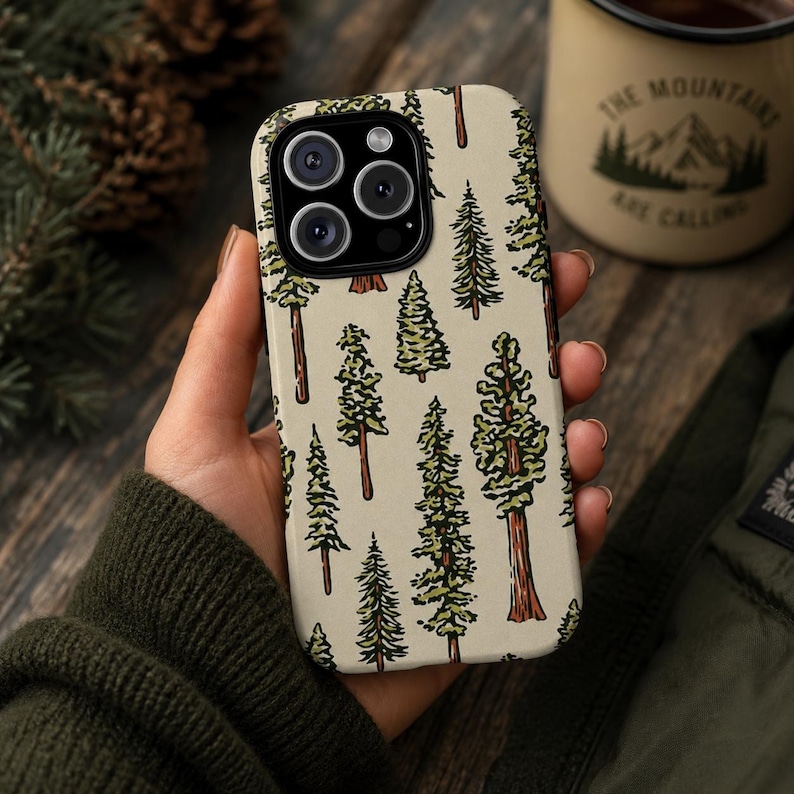 May include: A phone case featuring a repeating pattern of green and brown pine trees on a cream background. The phone case is held in a hand, with a green sweater sleeve visible. A mug with the text "The Mountains Are Calling" is in the background.