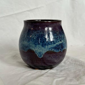 May include: A handcrafted ceramic vase with a rounded form. The vase displays a colour gradient, shifting from a deep purple at the base to a vibrant blue in the middle, and a darker purple at the rim. The glossy finish enhances the colours.