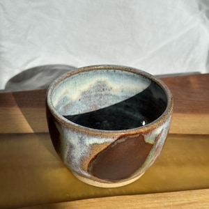 May include: A small, handmade ceramic bowl. The bowl has a rounded shape and a dark brown exterior with a lighter, mottled blue and white interior rim. It is likely used for serving or as a decorative piece.