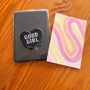 May include: A black phone case featuring a "GOOD GIRL" heart-shaped design and a card with abstract art in pink, orange, and yellow swirls. The items are on a wooden surface.
