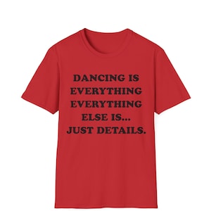 May include: A bright red short-sleeved t-shirt featuring black text printed on the front that reads: DANCING IS EVERYTHING EVERYTHING ELSE IS... JUST DETAILS. The garment has a crew neckline and a standard fit with short sleeves.