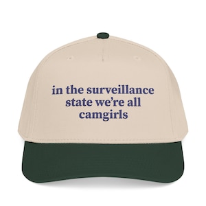 Puede incluir: Una gorra de béisbol de color beige con visera verde oscuro y un botón verde oscuro en la parte superior. La parte frontal de la gorra presenta el texto "in the surveillance state we're all camgirls" bordado en letras azul oscuro.